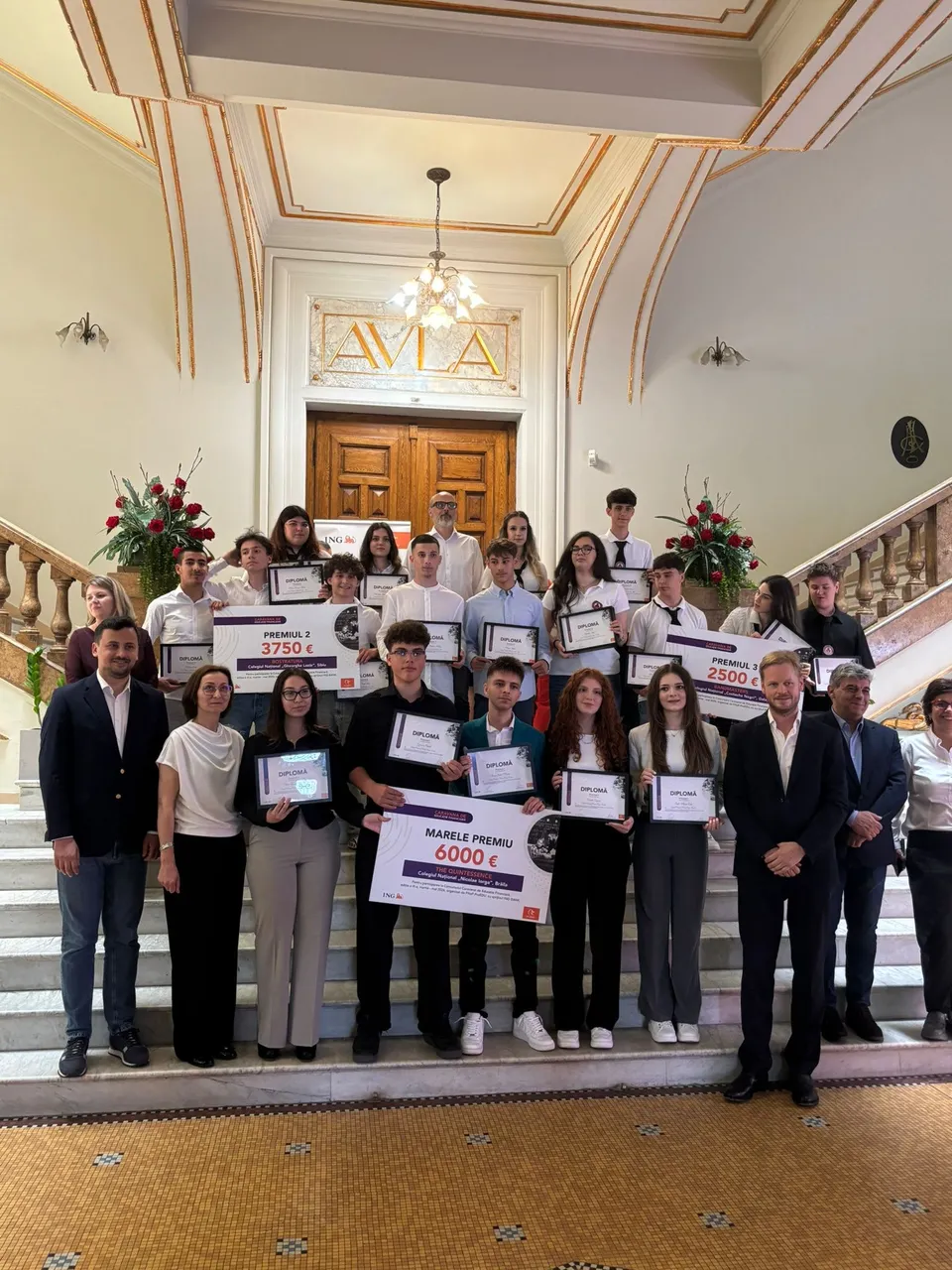 Un grup de oameni stă pe scări într-o încăpere interioară, mulți dintre ei ținând diplome și chei de premii de dimensiuni mari.
