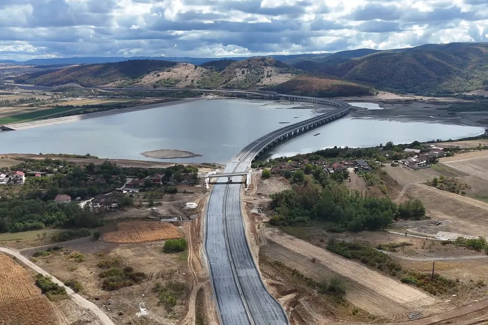 O porțiune de autostradă cu asfalt proaspăt se întinde printr-un peisaj rural, traversând un lac prin intermediul unui pod lung și curbat.