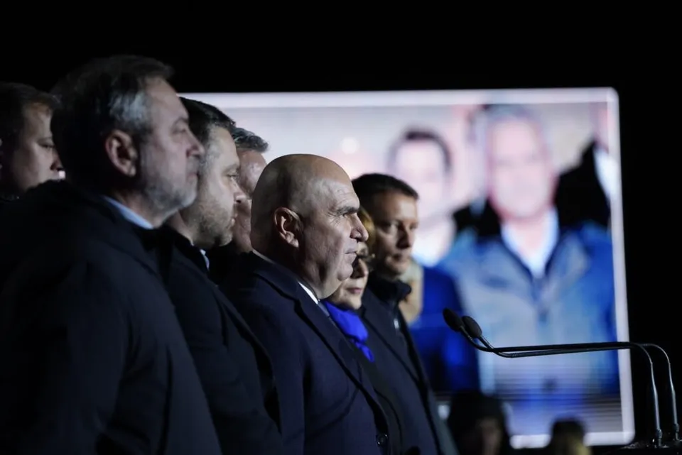 Un grup de bărbați în costume în plante de profil stau în fața unui ecran luminos care afișează imagini desfocate.