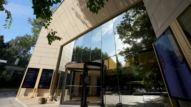 O clădire modernă cu fațadă de sticlă reflectă copacii verzi și cerul sub un unghi de jos.