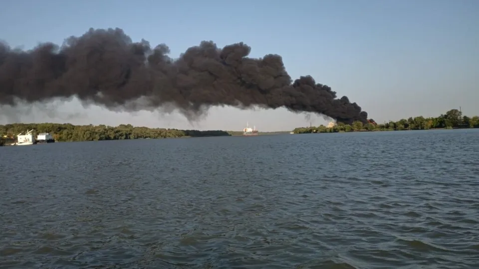 O imagine largă cu un fluviu sau lac în prim plan, cu un nor dens de fum negru care se ridică de pe țărmul din fundal, într-un peisaj cu arbori.