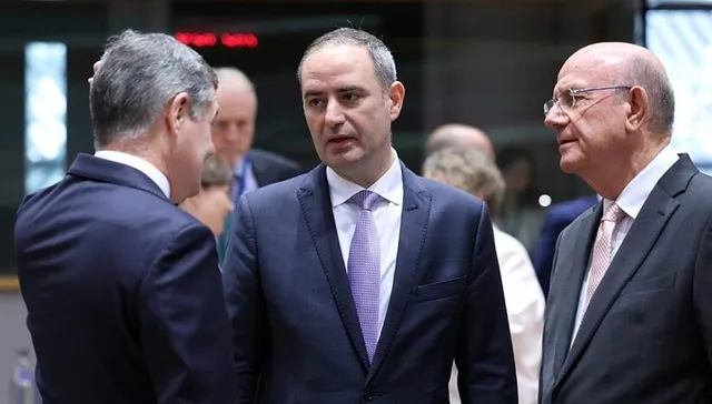 Trei bărbați în costume și cravate stau în picioare și discută într-un spațiu interior cu fundal desfocat.