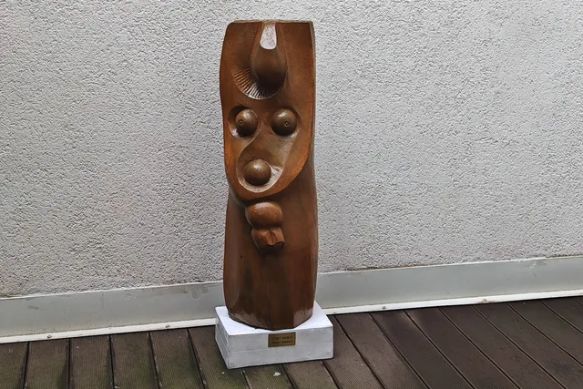 O sculptură verticală din lemn cu trăsături de față stilizate este așezată pe un soclu alb lângă un perete gri texturat.
