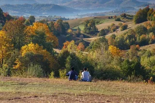 Două persoane stau pe o iarbă uscată, privind spre un peisaj format din dealuri, copaci cu frunziș colorat și munți în fundal.