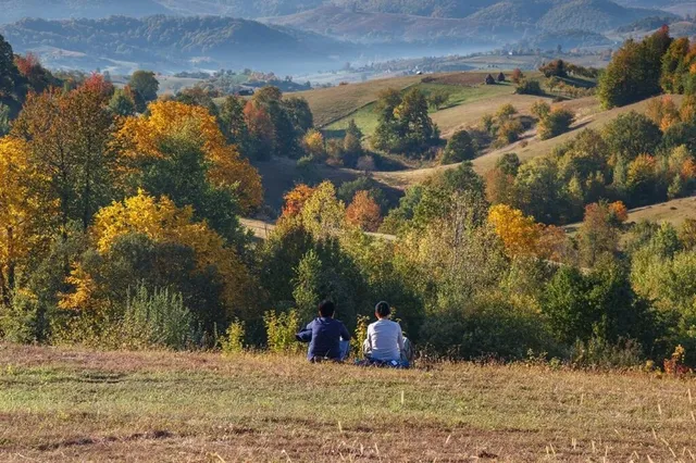 Două persoane stau pe o iarbă uscată, privind spre un peisaj format din dealuri, copaci cu frunziș colorat și munți în fundal.