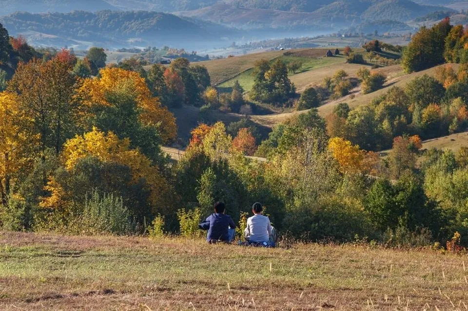 Două persoane stau pe o iarbă uscată, privind spre un peisaj format din dealuri, copaci cu frunziș colorat și munți în fundal.