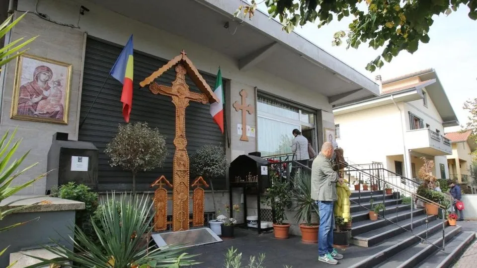 Un bărbat în sacou și pantaloni albaștri stă lângă scări, lângă o cruce mare din lemn și două steaguri pe fațada unei clădiri.