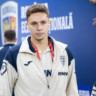 Un bărbat cu părul scurt și castan poartă o jachetă albă cu detalii albastre și un cordon roșu în jurul gâtului.