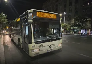 Un autobuz alb cu afișaj digital N103 circulă pe un drum asfaltat noaptea lângă un trotuar și clădiri urbane.