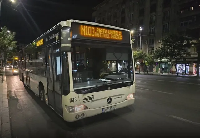 Un autobuz alb cu afișaj digital N103 circulă pe un drum asfaltat noaptea lângă un trotuar și clădiri urbane.