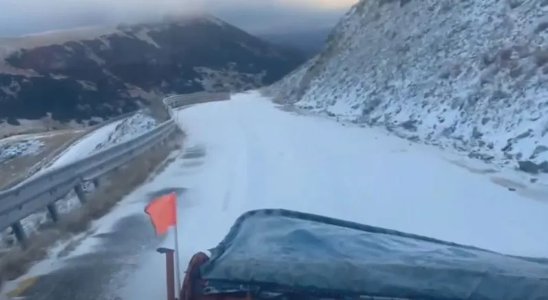O porțiune de drum montan acoperită de zăpadă este vizibilă de pe un vehicul, lângă un parapet metalic și un versant înzăpezit.