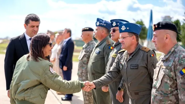 O femeie în jachetă verde strânge mâna unui bărbat în uniformă militară, în timp ce alți bărbați în uniforme și costume stau în fundal.