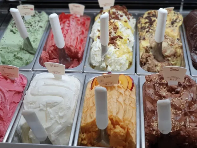 Mai multe tăvi metalice conțin diverse tipuri de înghețată colorată, fiecare având o spatulă cu mâner alb în interior.