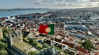 O vedere aeriană asupra unui oraș cu clădiri cu acoperișuri roșii, situat lângă un fluviu, cu un steag portughez agitat în prim-plan.