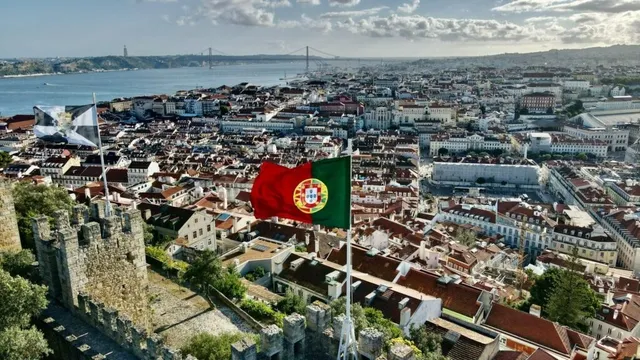 O vedere aeriană asupra unui oraș cu clădiri cu acoperișuri roșii, situat lângă un fluviu, cu un steag portughez agitat în prim-plan.