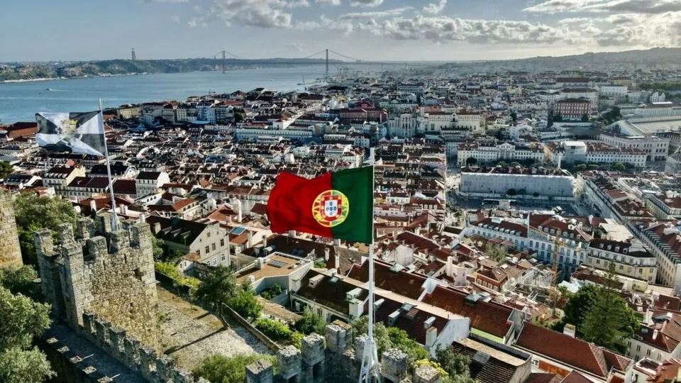 O vedere aeriană asupra unui oraș cu clădiri cu acoperișuri roșii, situat lângă un fluviu, cu un steag portughez agitat în prim-plan.