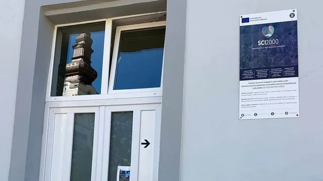 Un afiș albastru cu text alb și logo-uri este agățat pe un perete gri, lângă o ușă albă cu geamuri. Pe geam se reflectă o clădire. 