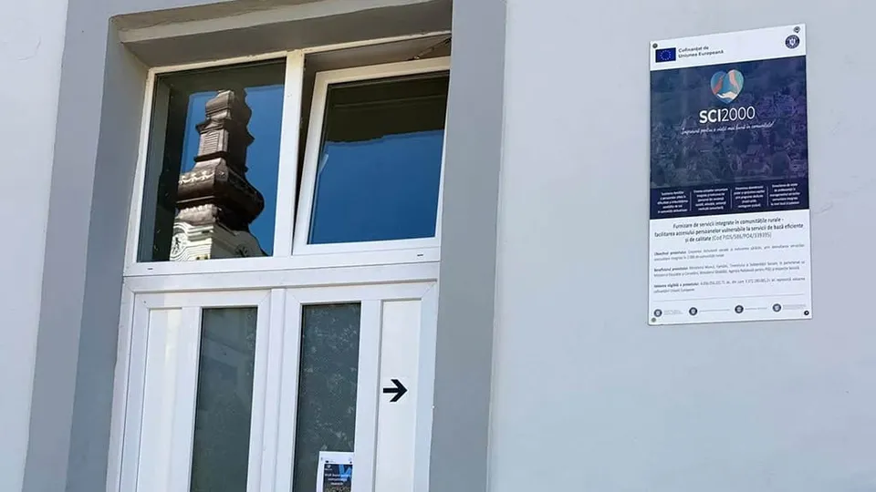 Un afiș albastru cu text alb și logo-uri este agățat pe un perete gri, lângă o ușă albă cu geamuri. Pe geam se reflectă o clădire. 
