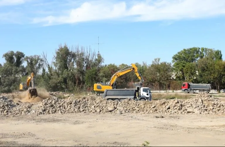 Două excavatoare galbene lucrează într-un șantier lângă două camioane cu benă, pe un teren cu grămezi de pământ și pietriș sub un cer senin.