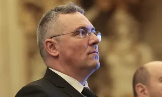 Un bărbat cu ochelari și costum negru privește în lateral într-un decor cu fundal neclar și lumină caldă.