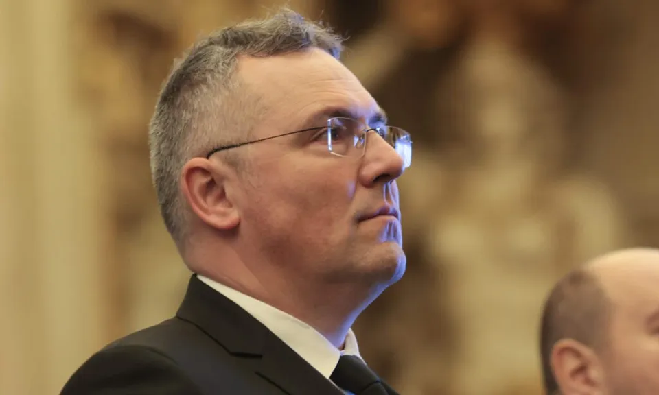 Un bărbat cu ochelari și păr scurt și cărunt poartă un costum negru și o cămașă albă, privind spre lateral.