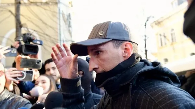 Un bărbat cu șapcă și jachetă neagră ridică mâna stângă în timp ce este înconjurat de microfoane și telefoane mobile.