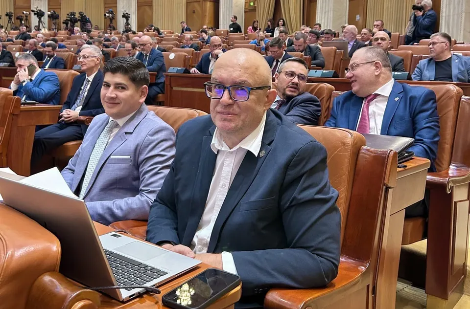 Un grup de bărbați în costume oficiale stau așezați în scaune de piele maro într-o sală de conferințe cu birouri de lemn.