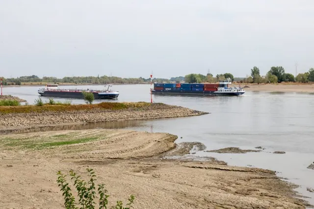 Două nave de transport navighează pe un râu larg lângă o țărm nisipos și uscat sub un cer înnorat.