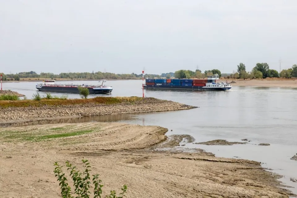 Două nave de transport navighează pe un râu larg lângă o țărm nisipos și uscat sub un cer înnorat.