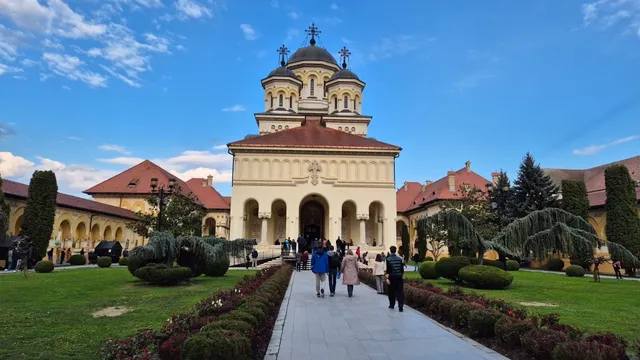 O catedrală cu cupole și turle se ridică sub un cer albastru, înconjurată de un spațiu verde cu alei și persoane care merg pe drumuri pietruite.