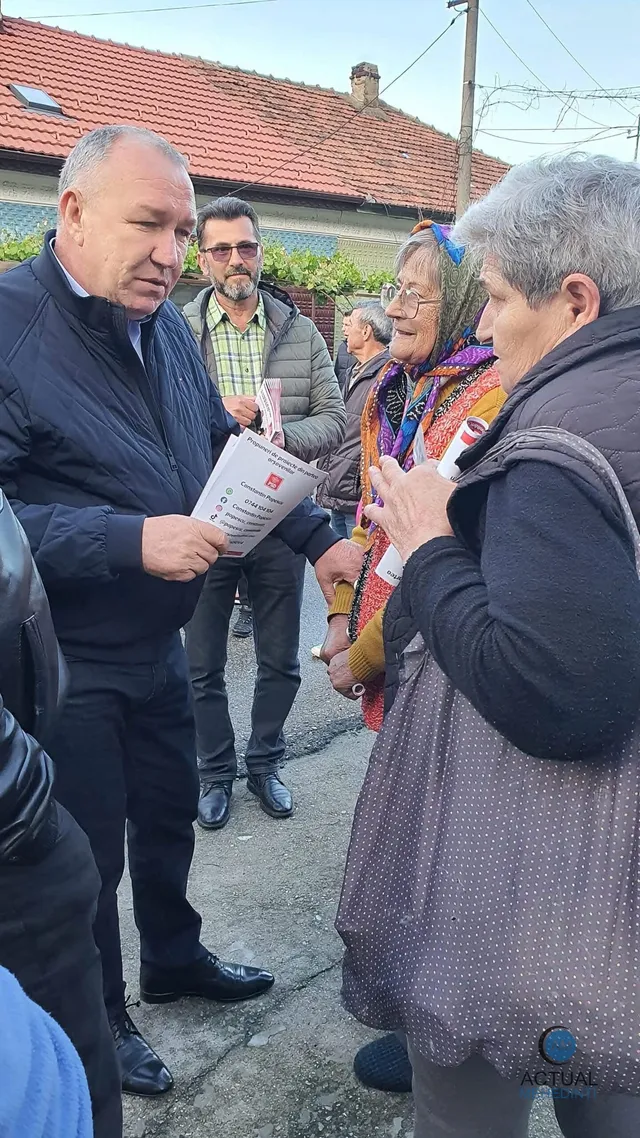 Un bărbat într-o jachetă neagră ține o broșură albă în timp ce stă în fața unui grup de persoane pe o stradă.