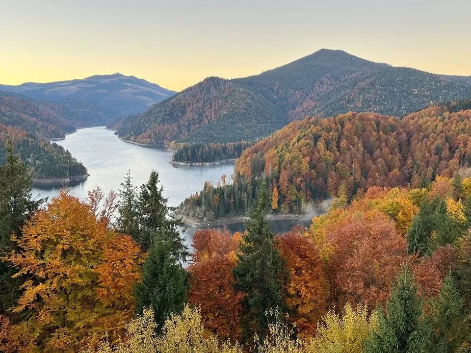 Un lac se află între dealuri acoperite de păduri cu copaci în nuanțe de portocaliu, galben și verde sub un cer deschis.