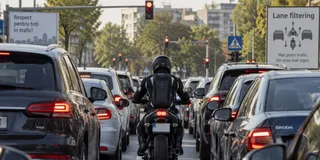 Un motociclist cu cască neagră și vestă de protecție se află între rânduri de mașini în trafic pe un drum urban.