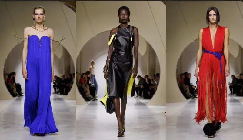 Trei modele merg pe o pistă de catwalk purtând rochii lungi de culori albastru, negru și roșu în fața unui public așezat.