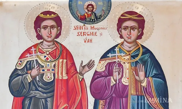 Două figuri pictate în stil iconografic poartă haine colorate și țin cruci mici în mâinile drepte pe un fundal deschis cu haloane circulare.