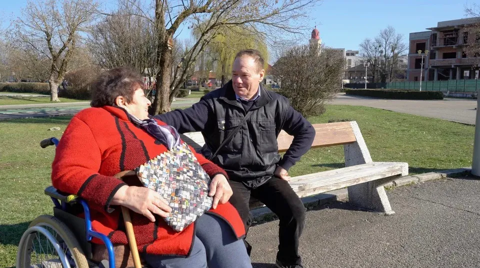 Un bărbat în haine negre stă pe o bancă de lemn lângă o femeie cu o haină roșie așezată într-un scaun cu rotile.