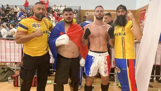 Patru bărbați stau în picioare într-o sală de sport, doi purtând tricouri galbene și doi fiind torți sau sportivi cu echipament de luptă.