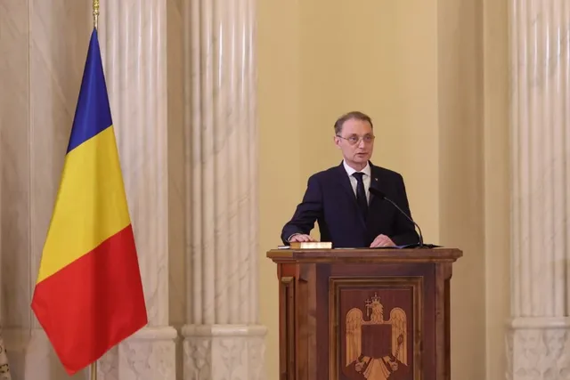 Un bărbat cu ochelari și costum închis stă în spatele unui podium de lemn decorat cu un blazon, lângă un steag tricolor.