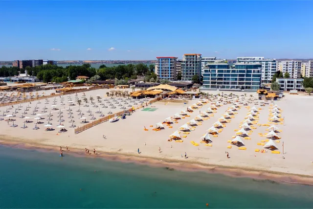 O vedere aeriană a unei plaje cu nisip deschisă, cu numeroase șezlonguri și umbrele sub parasole albe și galbene, situată în fața unor clădiri moderne.