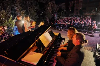 Trei muzicieni cântă pe o scenă cu un pian de concert, în fața unui public așezat pe scaune în aer liber noaptea.