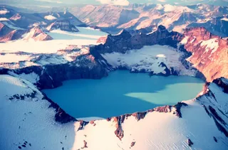 O vedere aeriană a unor munți acoperiți de zăpadă și un lac albastru situat într-un crater montan.