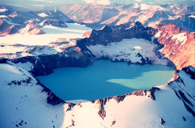 O vedere aeriană a unor munți acoperiți de zăpadă și un lac albastru situat într-un crater montan.