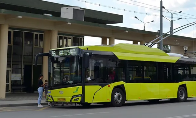 Un autobuz electric de culoare galbenă este oprit în fața unei clădiri cu coloane lângă un trotuar unde stă o persoană.