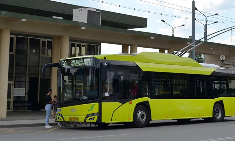 Un autobuz electric de culoare galbenă este oprit în fața unei clădiri cu coloane lângă un trotuar unde stă o persoană.