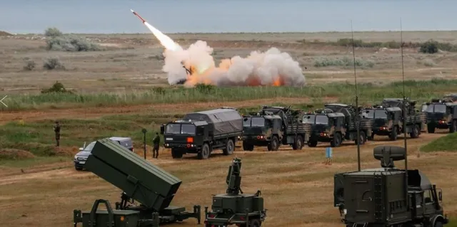 Mai multe vehicule militare verzi sunt parcate pe un teren deschis, în timp ce un proiectil este lansat din fundal, lăsând o urmă de fum.