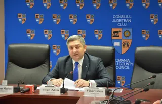 Un bărbat cu sacou închis și cravată albastră stă la o masă de conferințe în fața unui panou albastru cu embleme.