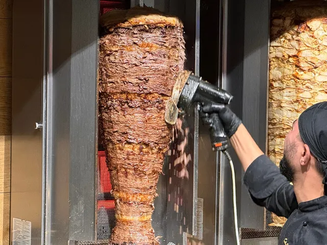 Un bărbat cu căciulă neagră și mănuși folosește un aparat electric pentru a tăia feliile de carne de pe un rotisor vertical.