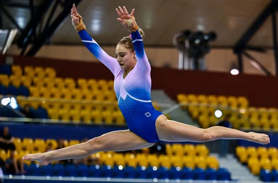 O gimnastă într-o sală de gimnastică cu fundalul de scaune galben și albastru, arătând o poziție de split aerian cu brațele ridicate.