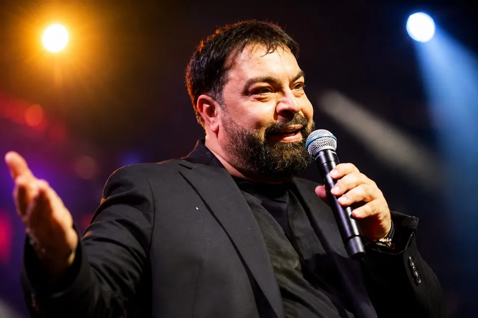 Un bărbat cu barbă și păr scurt, îmbrăcat în sacou negru, ține un microfon argintiu și negru în mână în fața unor lumini colorate.