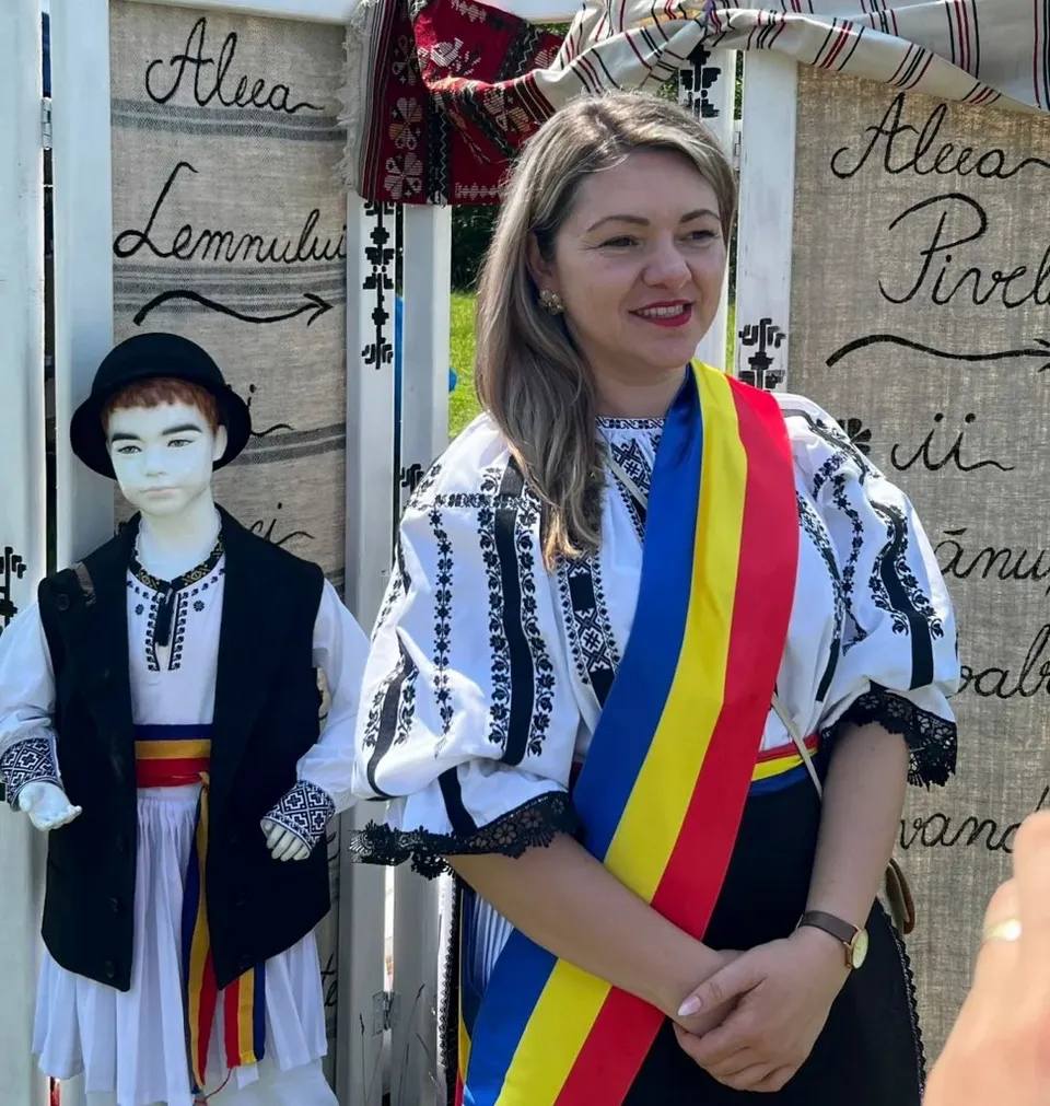 O femeie zâmbinde poartă o cămașă tradițională albă cu broderii negre și o eșarfă tricoloră pe umăr, stând lângă un manechin îmbrăcat în costum popular.